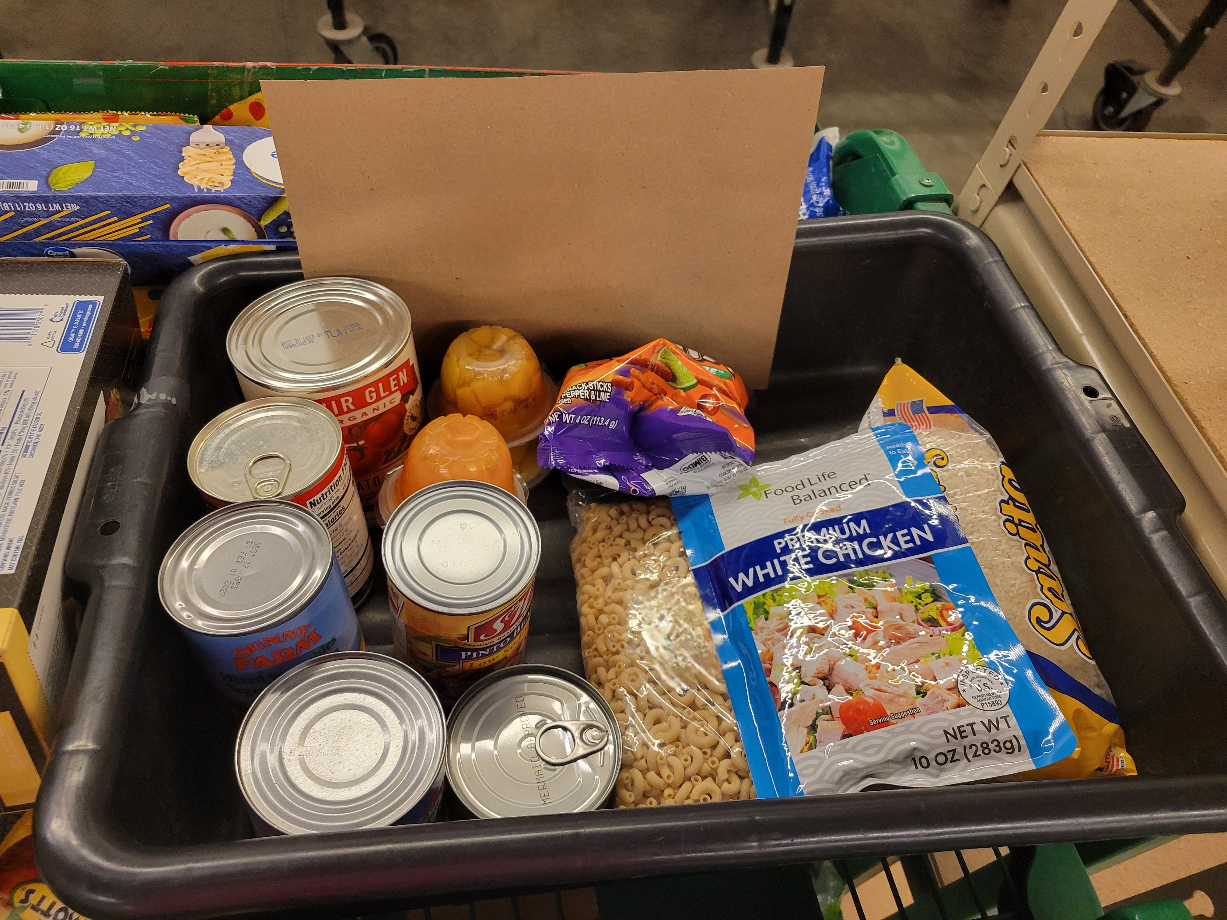 Small black plastic container filled with canned corn, canned tuna, canned tomatoes, canned mixed vegetables, four cups of mandarin oranges, bag of rice, pouch of chicken,  bag of elbow macaroni, and small bag of spicy chips.