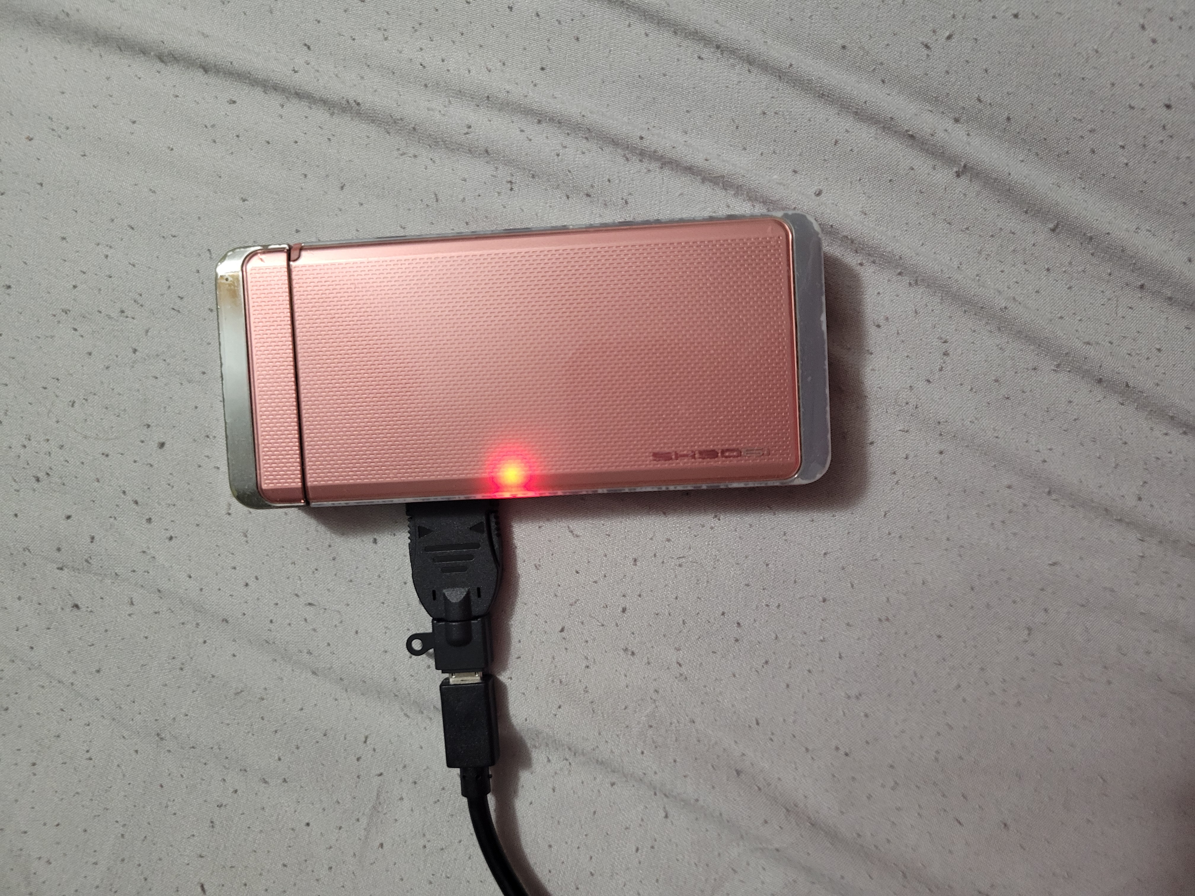 Pink flip phone with a red light on and a charger converter sticking out on the side.