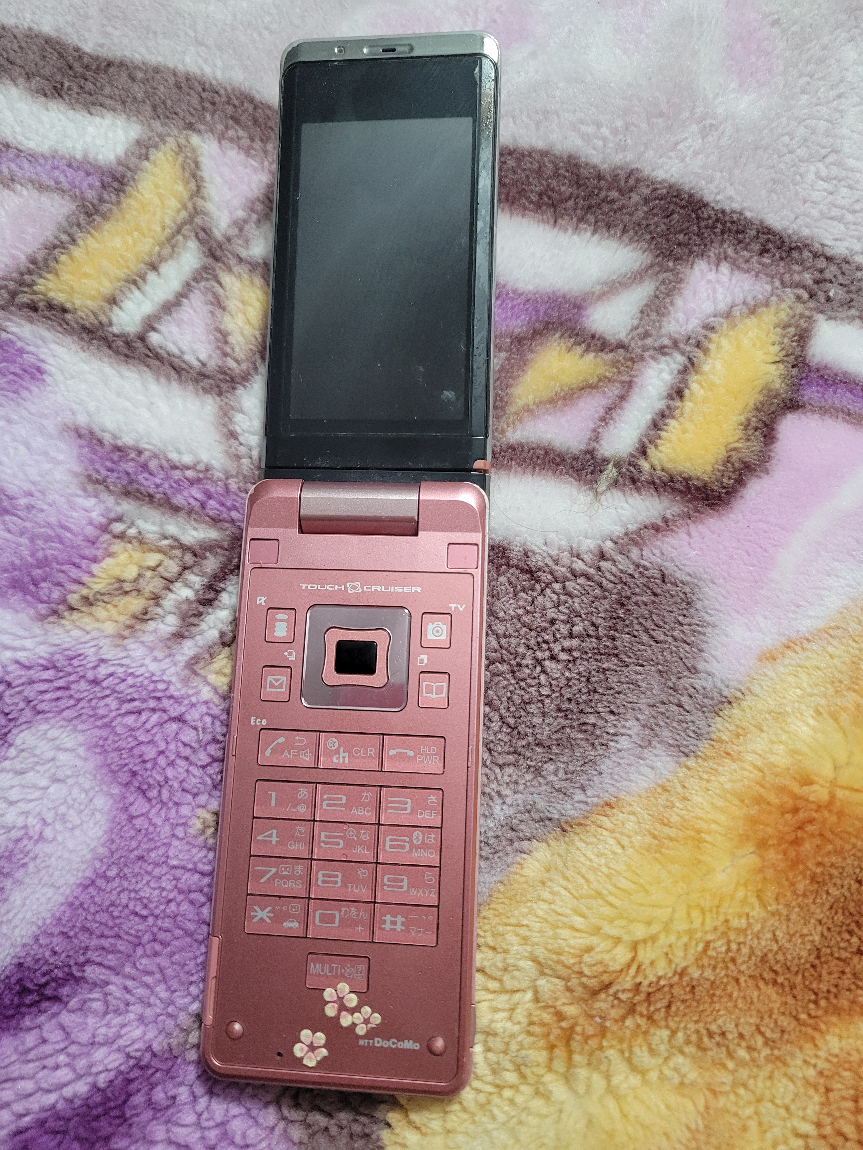 A open pink Japanese flip phone with small pink flowers on the bottom placed on top of a fuzzy blanket